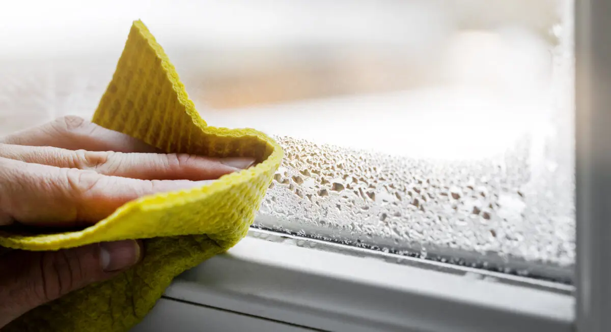 Close-up of a hand using a yellow cloth to wipe condensation from a window, showing water droplets on the glass and window frame.