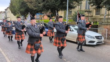 The East Riding Pipe Band dressed in grey jackets and hats with orange kilts playing their pipes while walking down the road.