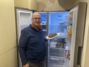 Terry Andrews, Swanky Village Hall Chairman, stands in from of on opened fridge freezer holding a plate of food.