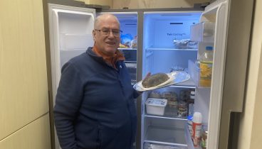 Terry Andrews, Swanky Village Hall Chairman, stands in from of on opened fridge freezer holding a plate of food.